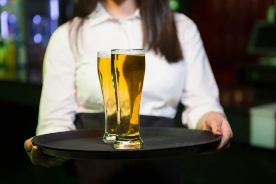person dressed in a white button-down shirt holding a black tray with two tall glasses of beer