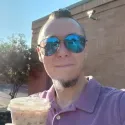 image of Brian McQuay, with short brown hair and goatee, in a mauve polo smiling at the camera holding an iced coffee and wearing reflective blue aviator sunglasses