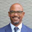 headshot of Will Lukela, wearing a blue suit, white button-down shirt and patterned blue tie, with a black and grey bearded goatee and angular black glasses