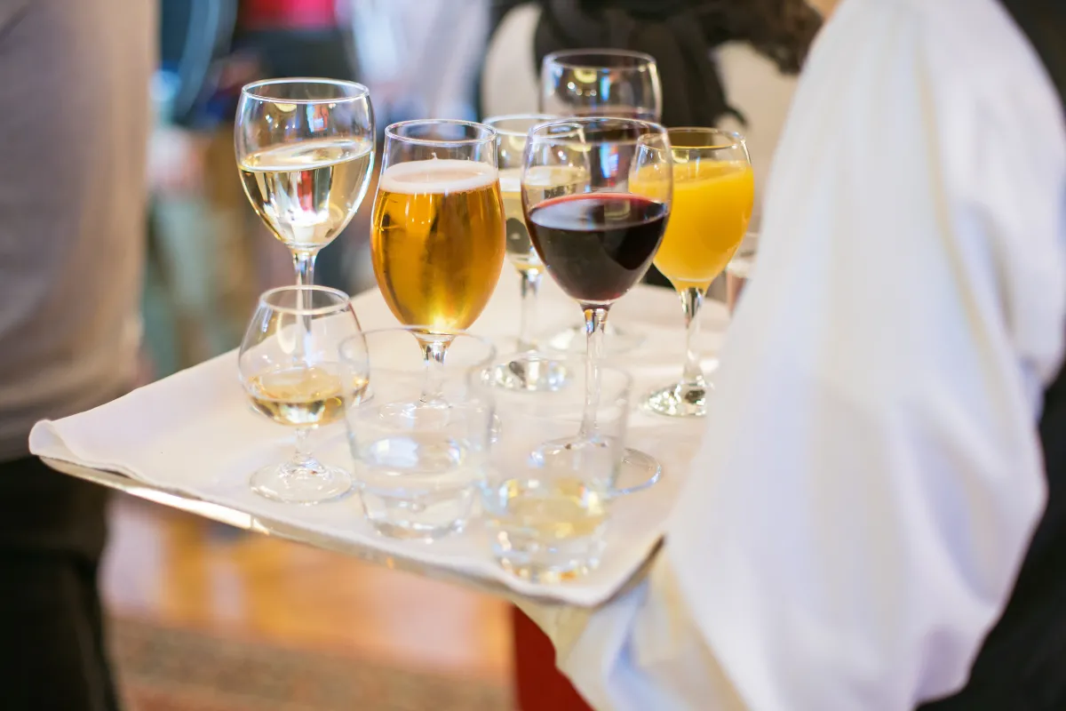 image of a person in a white button-down shirt and black vest holding a white tray with a variety of glasses, including wine, a cocktail, champagne and orange juice