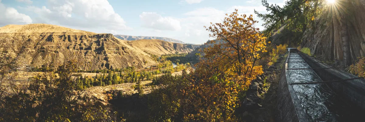 a panorama of a bright sunny day overlooking a large hill on the left and some orange, yellow and green trees and running water on the right