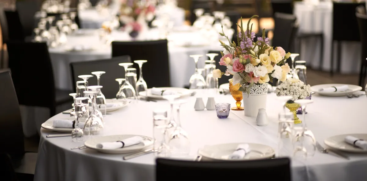 A round banquet dinner table with a white tablecloth and place settings, with two glasses per plate and a bouquet of flowers in the center