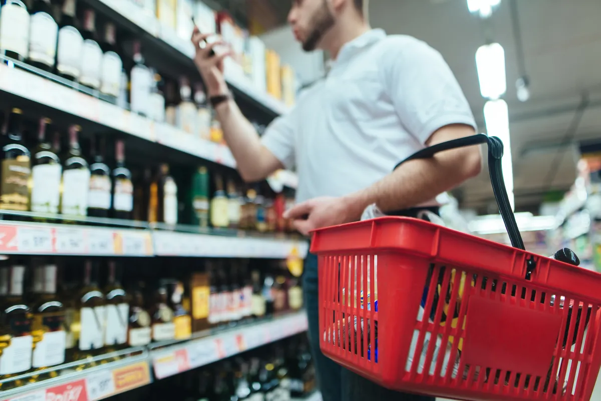 person in a grocery store, wearing a light polo and holding a red shopping basket, examining wine bottles on a grocery store shelf