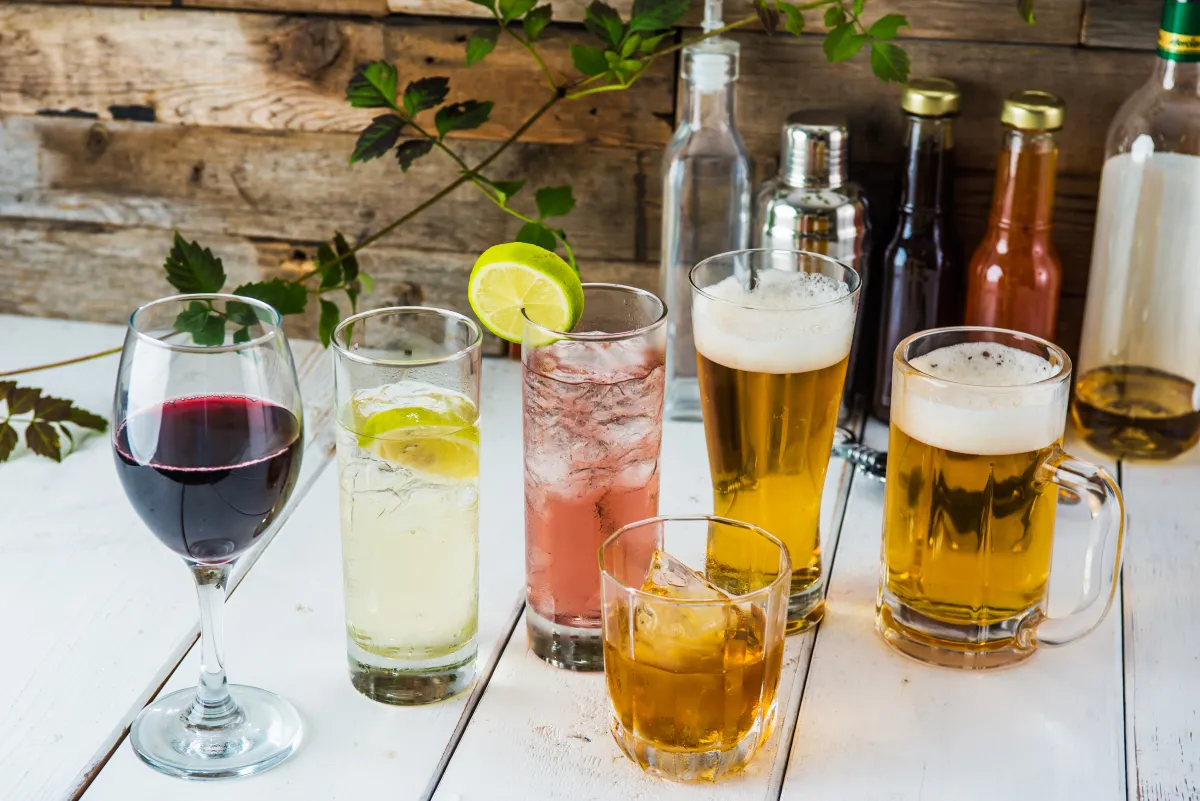 multiple drink glasses lined on a wooden table, red wine, cocktails, beer, with bottles in the background