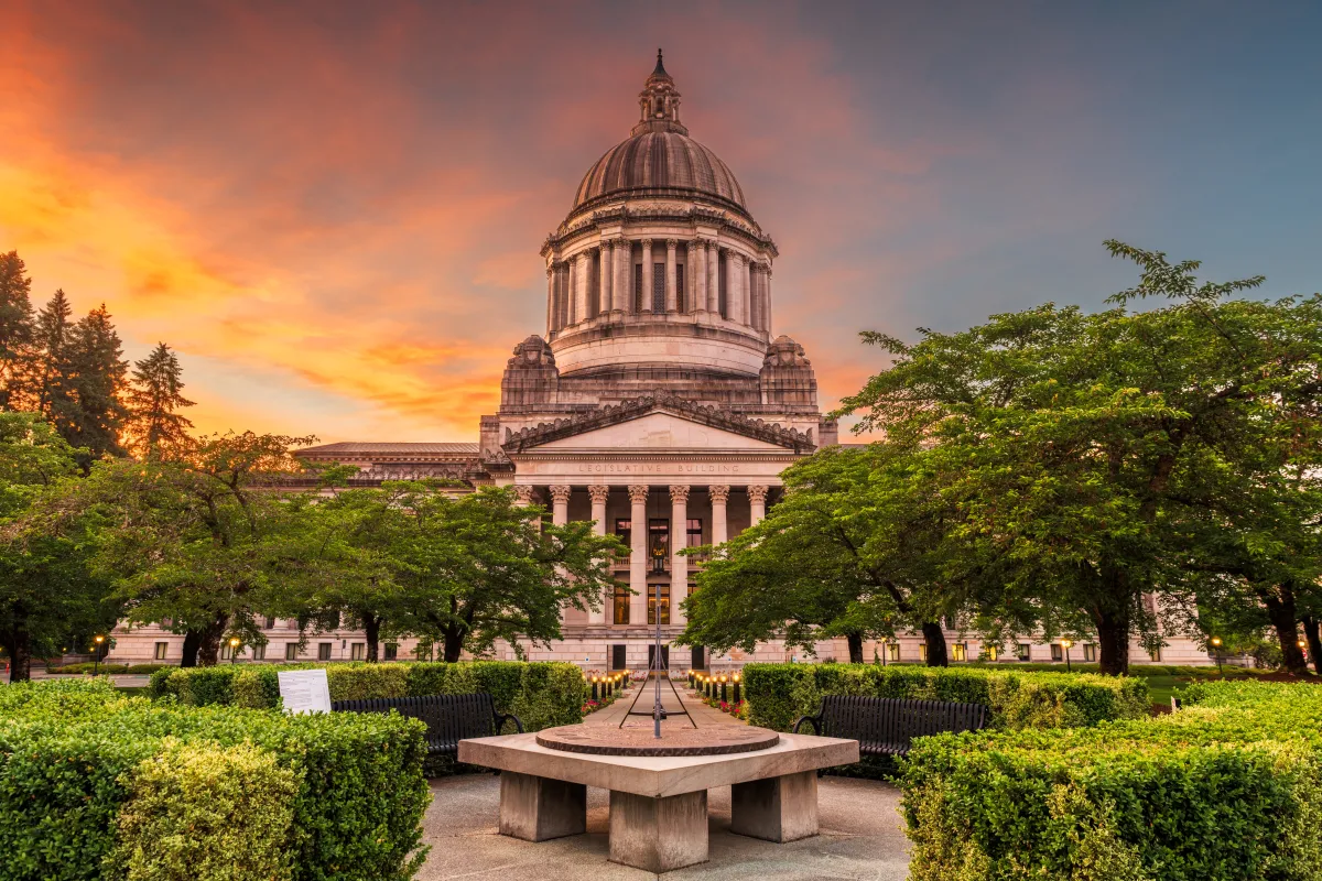 image of the Olympia capitol building and campus, the sunset turning the sky shades of orange, pink and blue with greenery surrounding the fountain in front of the building