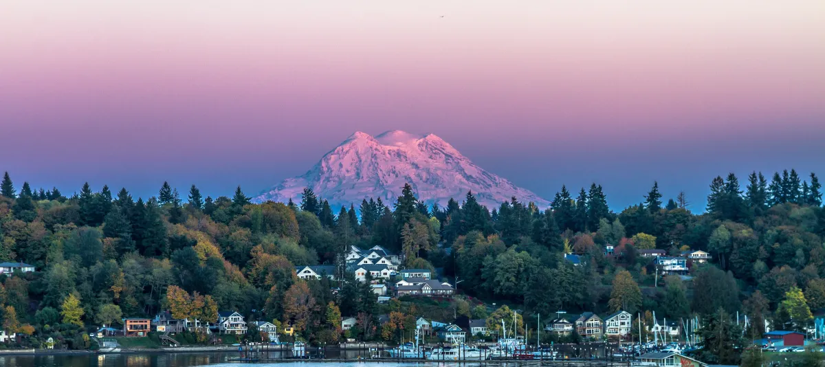 image of Mount Rainier at sunset, the sky a rainbow of blues, pinks, oranges and yellow, with trees and houses overlooking the south sound at the base of the mountain