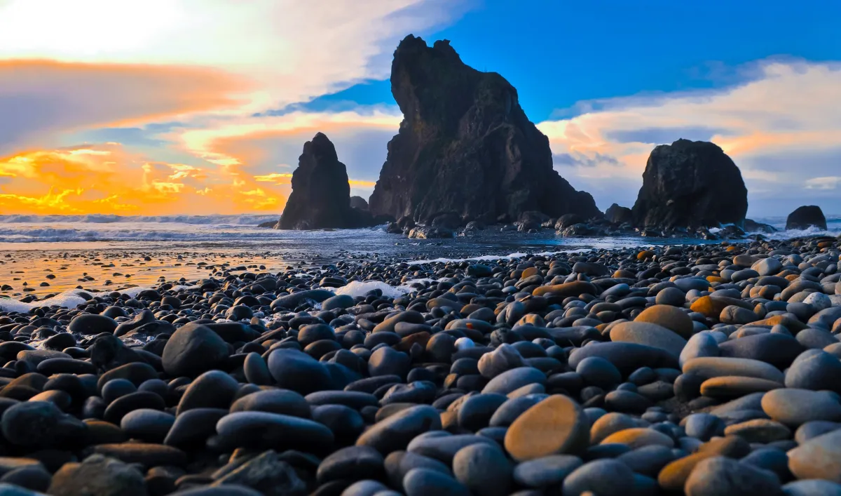 a rocky pebbled beach with smooth waves crashing over the smooth stones, three large rocky structures reaching up in the distance, a sunset behind the water turning the sky shades of blue, orange and white