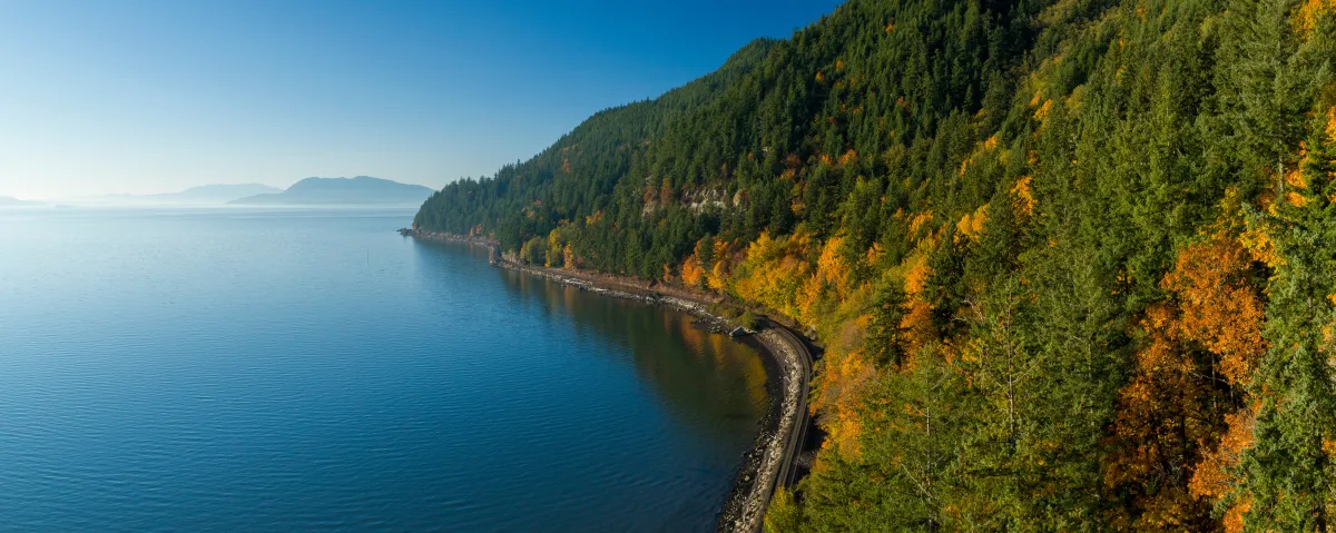 blue open water with a clear sky, a highway rounding rolling hills covered in green, yellow and orange trees on the right side