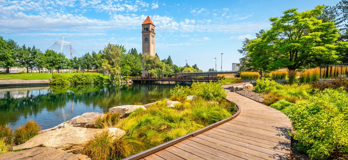 Image of the Spokane riverfront on a sunny day, a boardwalk weaving along the waterfront surrounded by greenery and a clock tower reaching into a blue sky