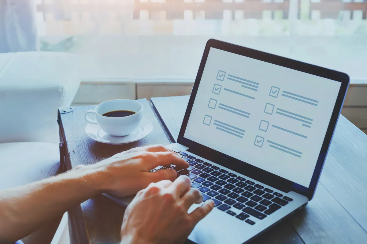 person sitting at a table with a white coffee mug and saucer full of coffee typing on a laptop showing a screen with checkboxes and light grey lines