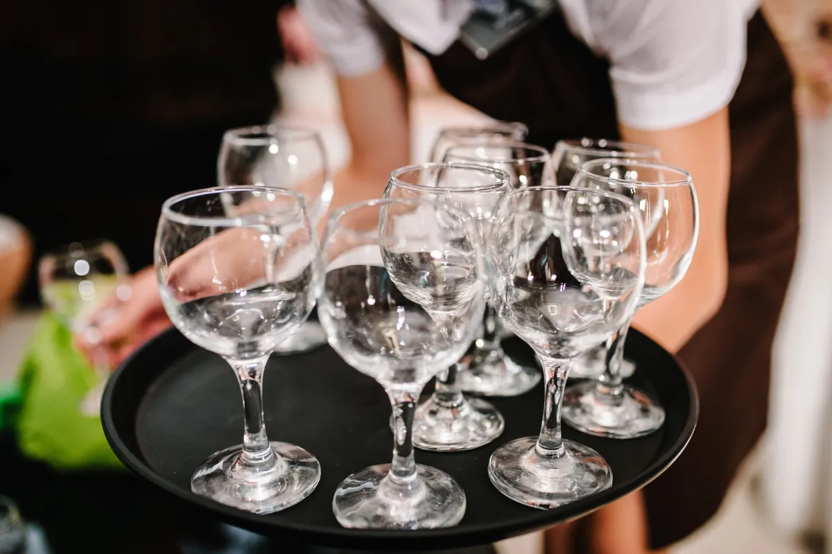 person in a black apron and white short-sleeved shirt holding a black tray full of empty wine glasses