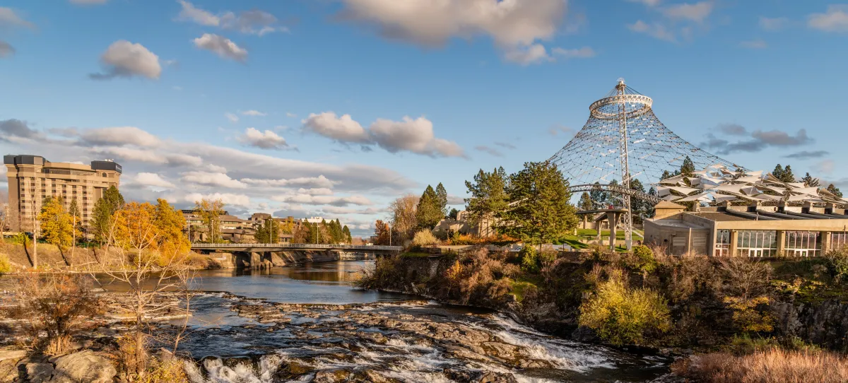 landscape image of Spokane, Washington with a river, buildings, and tall metal structure with the background of a blue sky and some clouds