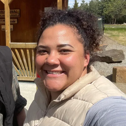 Officer Tiffinni Archie smiling, standing in front of a wooden building wearing a beige puffer vest and light blue shirt