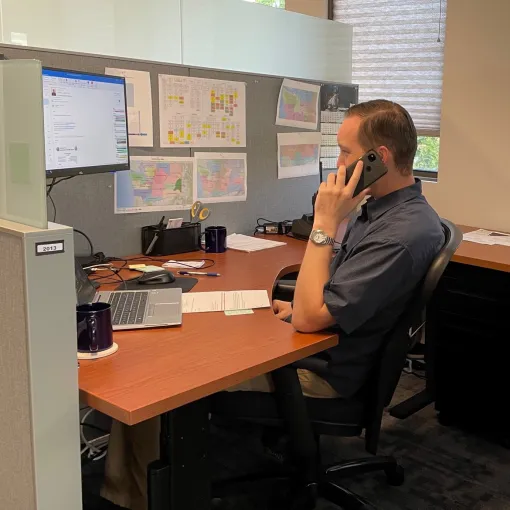 Lieutenant E.P. Hackenberg in a dark grey polo, sitting at a cubicle desk speaking on their cell phone