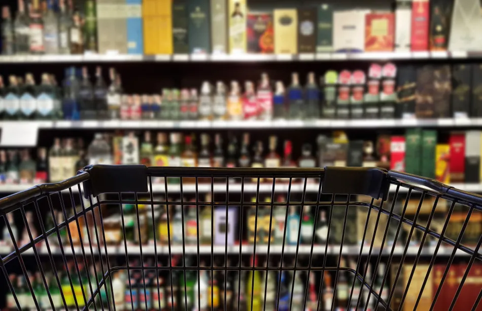image of a black grocery cart in focus in front of a blurred aisle of shelves containing bottles of liquor